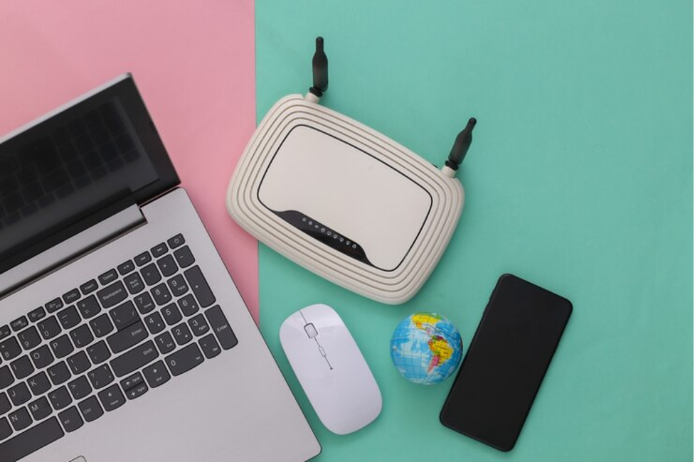 Laptop silver di samping router putih, mouse nirkabel, dan smartphone, semuanya diletakkan di atas latar berwarna pink dan hijau, sebagai ilustrasi untuk membahas harga modem wifi.